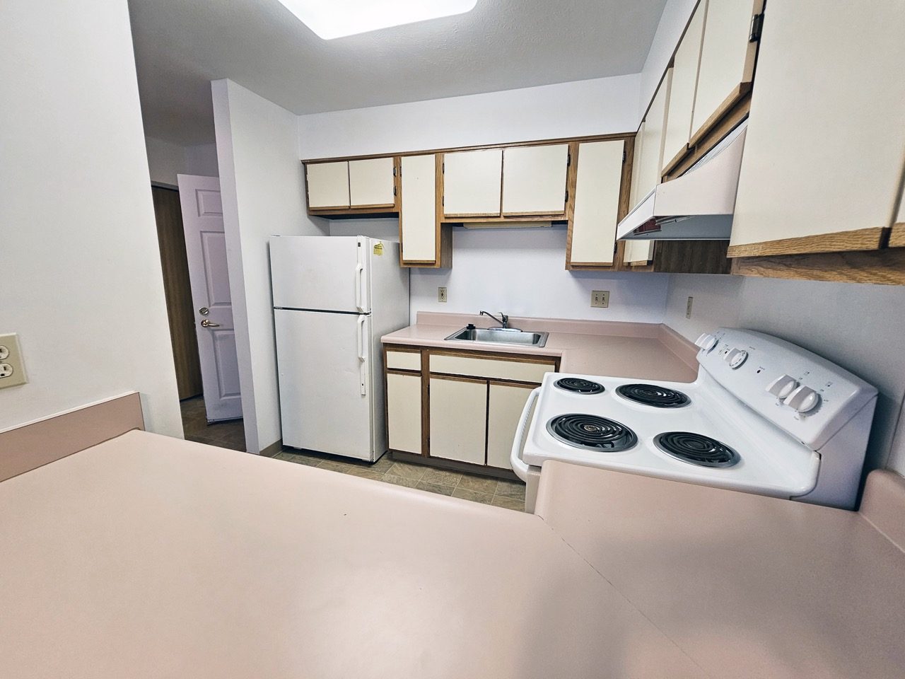 Dated apartment kitchen with original wood-tone cabinets, white range and worn vinyl flooring before renovation.